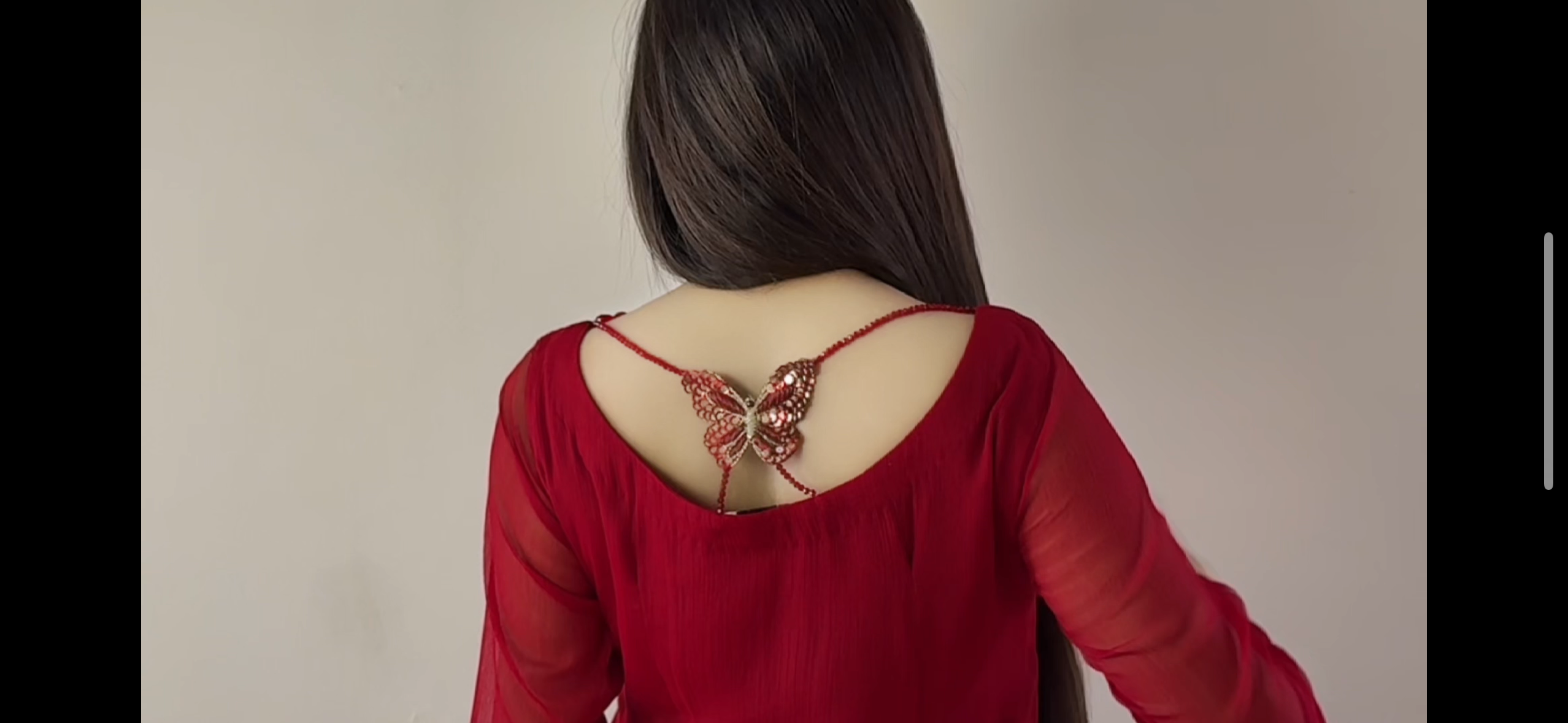 A person wearing a sheer red long-sleeve top, shown from the back with dark hair, featuring a delicate red chain accessory with a butterfly embellishment at the open back, set against a plain light gray background.